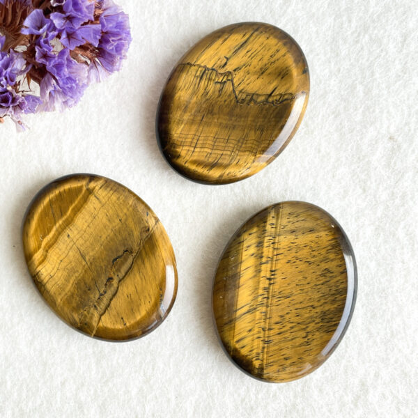 Three oval tiger's eye gemstones with bands of golden and brown colors displayed on a white textured background alongside dried purple flowers.