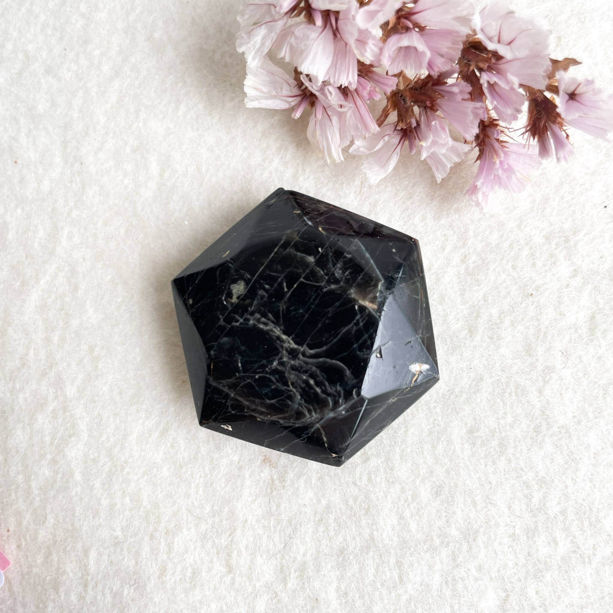 A polished black hexagonal gemstone with visible white veining lies on a textured white surface, accompanied by pink cherry blossom petals nearby.