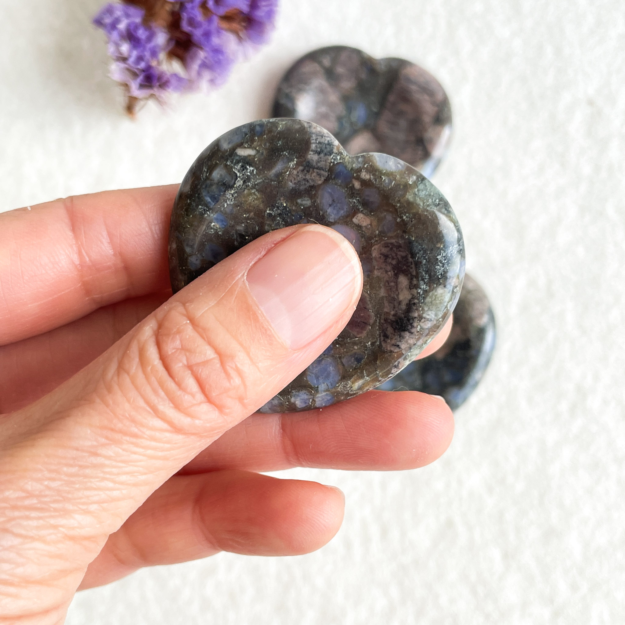 A hand holding a polished heart-shaped stone with a mix of dark and light blue colors, with similar stones and purple dried flowers visible in the background on a white surface.