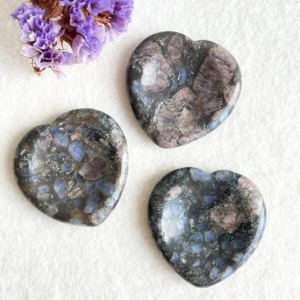 Two polished heart-shaped stones with mottled blue and brown patterns on a light background, accompanied by a small bundle of dried purple flowers on the upper left side.