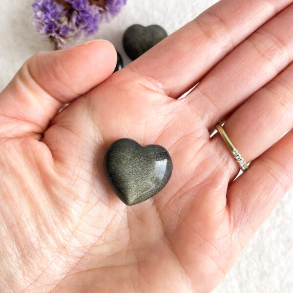 A person's palm holding a polished heart-shaped stone, with a glimpse of a diamond ring on their finger and a blurred background of purple flowers and another heart-shaped object.