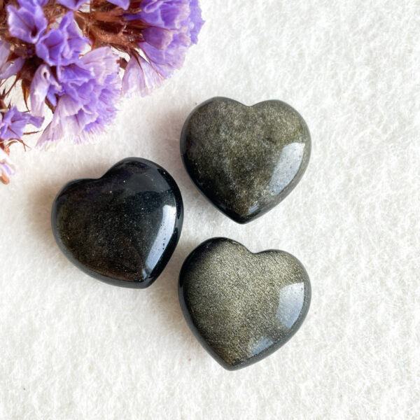 Two polished, heart-shaped stones on a textured white background, with a corner of purple flowers visible in the top left.