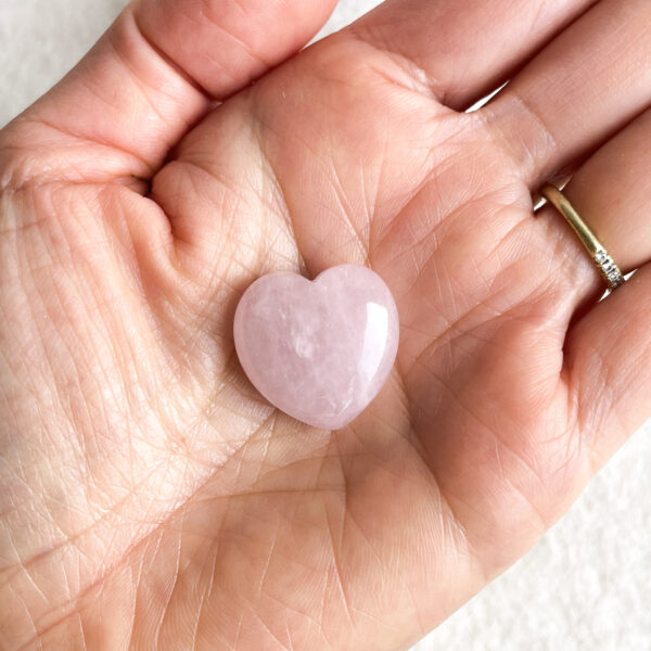 A hand holding a polished rose quartz heart with a gold ring on the ring finger.
