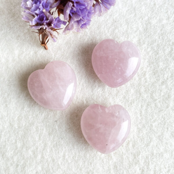 Two polished rose quartz stones shaped like hearts on a white textured background with dried purple flowers partially visible at the top left corner.