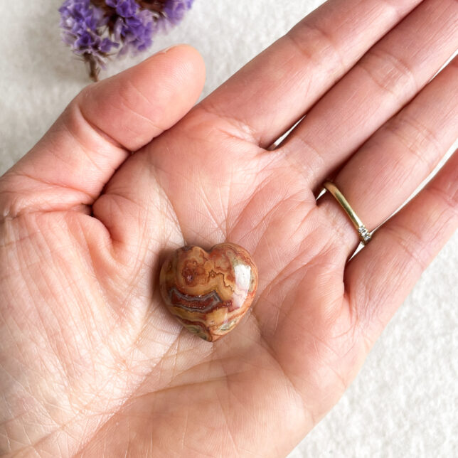 A person's hand with visible ring on the ring finger, holding a heart-shaped multicolored stone with purple flowers blurred in the background.