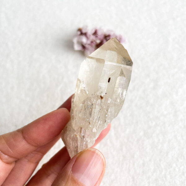 A close-up of a person's fingers holding a translucent quartz crystal against a white background, with a small purple flower out of focus in the background.