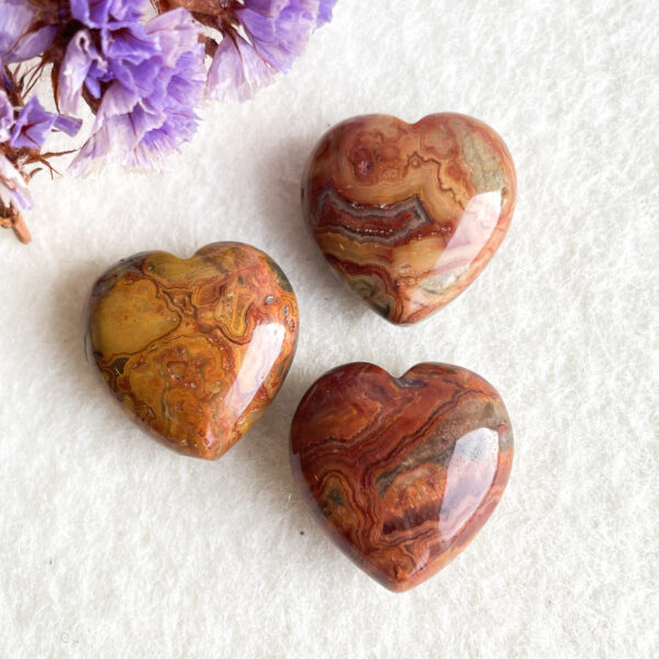 Three polished, heart-shaped stones with layered earthy red, brown, and cream colors on a white background, accompanied by dried purple flowers in the upper left corner.