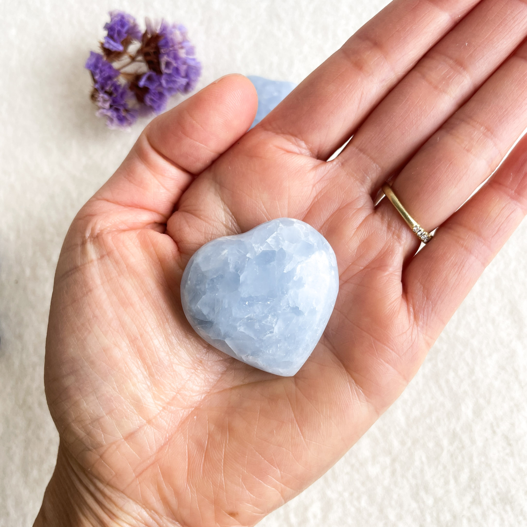 A person's open hand holding a heart-shaped blue crystal, with a gold ring on their ring finger, and a small bunch of dried purple flowers in the upper left corner against a textured off-white background.
