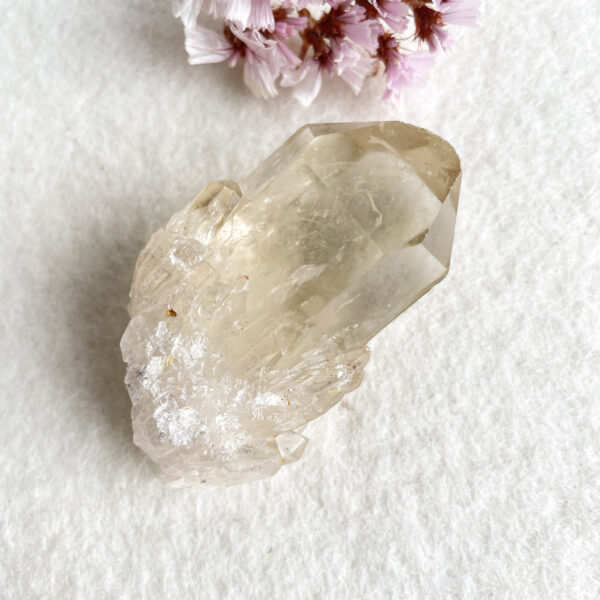 A large, translucent quartz crystal with multiple facets resting on a white textured surface, with blurry pink flowers in the background on the upper left corner.