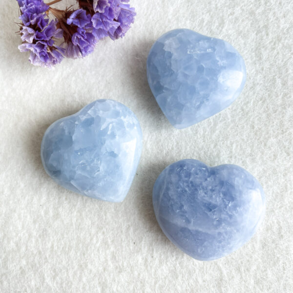 Three blue crystal hearts on a white background with a small bunch of dried purple flowers in the top left corner.