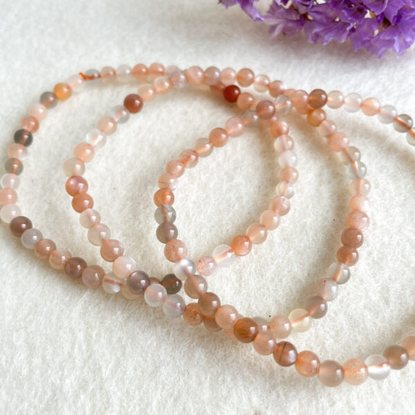 Three strands of translucent beaded bracelets in shades of pink and brown curling loosely on a white and textured surface with purple flowers in the corner.