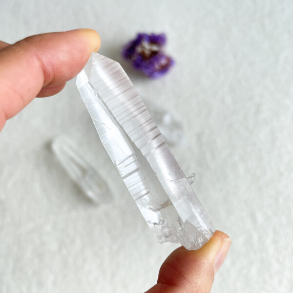 A person's fingers holding a clear quartz crystal with a blurred purple flower and another crystal in the background.