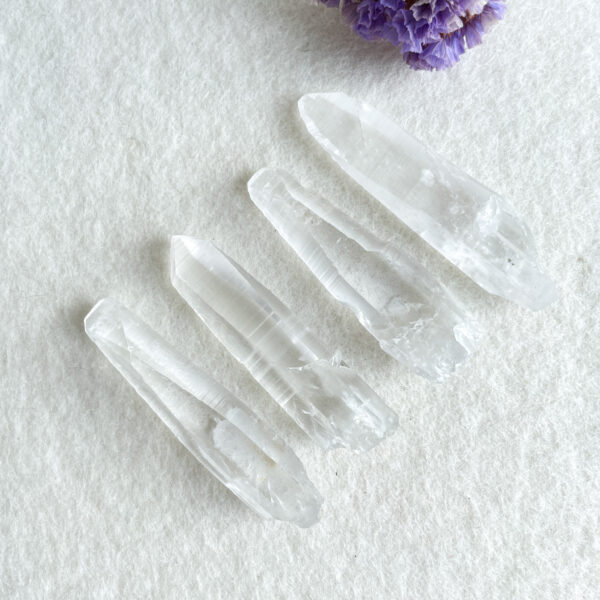 Four clear quartz crystals arranged on a white textured background with a purple flower in the top right corner.