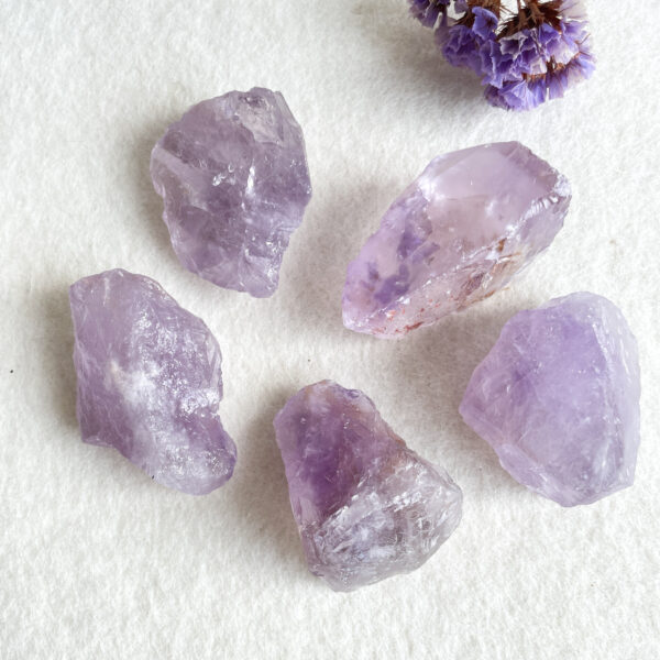 Five raw amethyst crystals displayed on a white background, with a cluster of purple dried flowers partially visible at the top right corner.