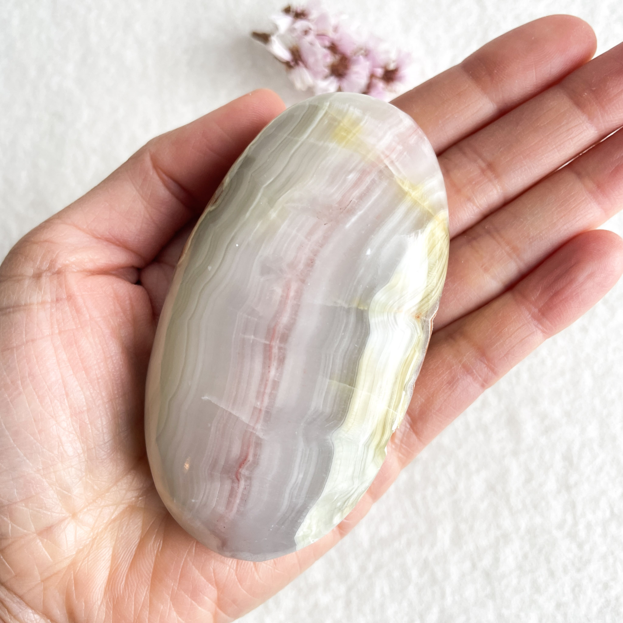 A hand holding a polished oval onyx stone with white, gray, and yellow banding patterns, against a white background with a blurred small pink flower in the corner.