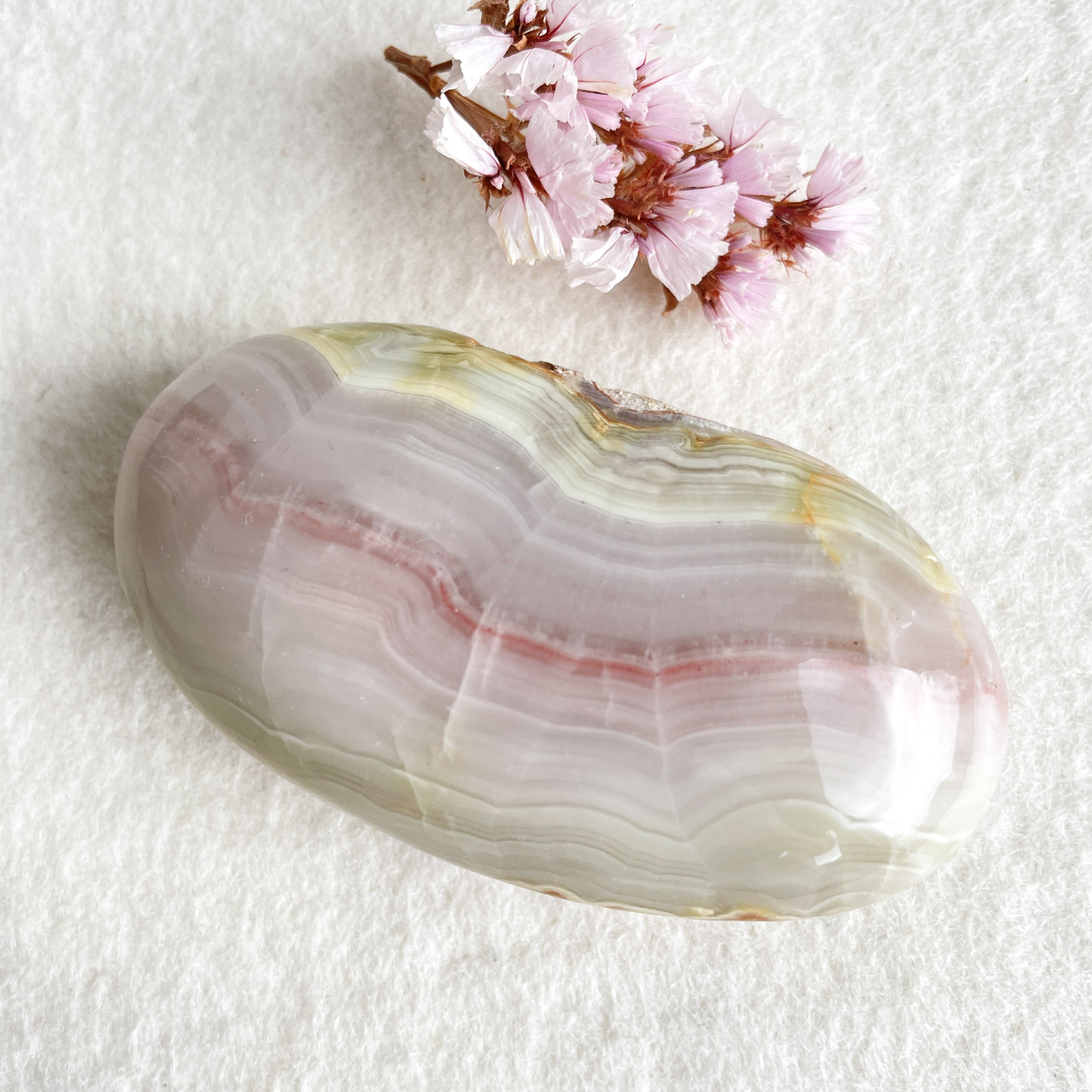 A polished oval agate stone with translucent layers of pink and white, alongside a few sprigs of pink cherry blossoms, on a white textured background.