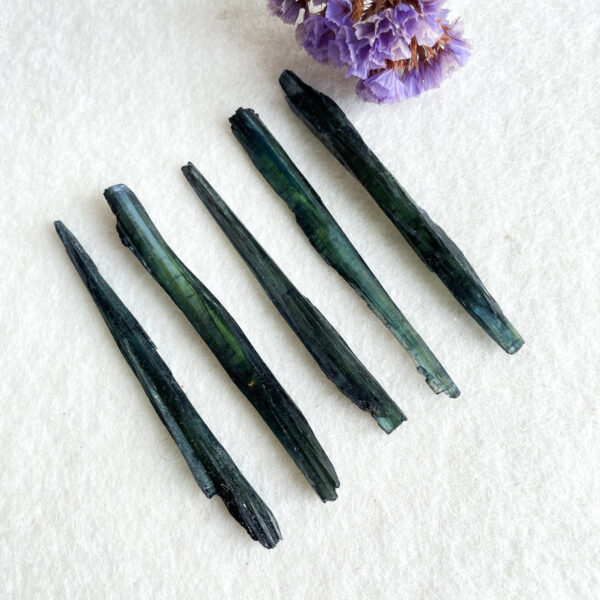 Five dark green tourmaline crystal sticks arranged parallel to each other on a white textured surface with a cluster of dried purple flowers in the upper right corner.