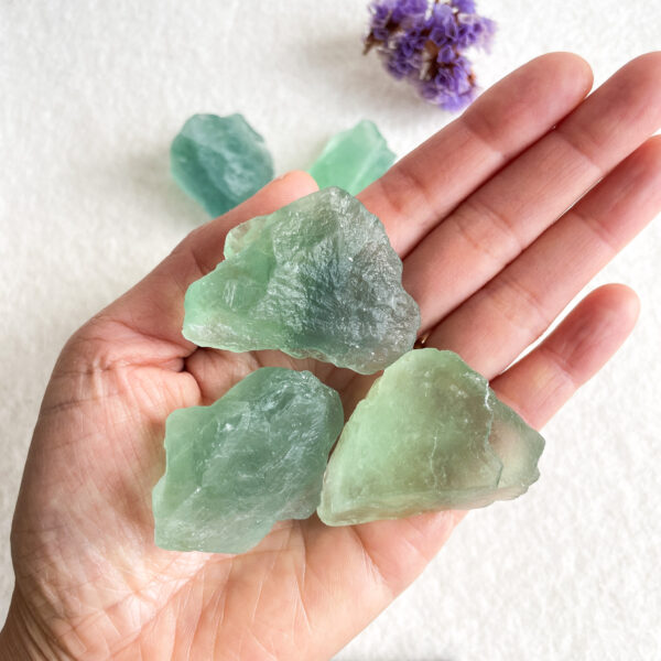 A hand holding three chunks of raw green fluorite crystals against a white textured background, with a hint of purple flowers visible in the top corner.