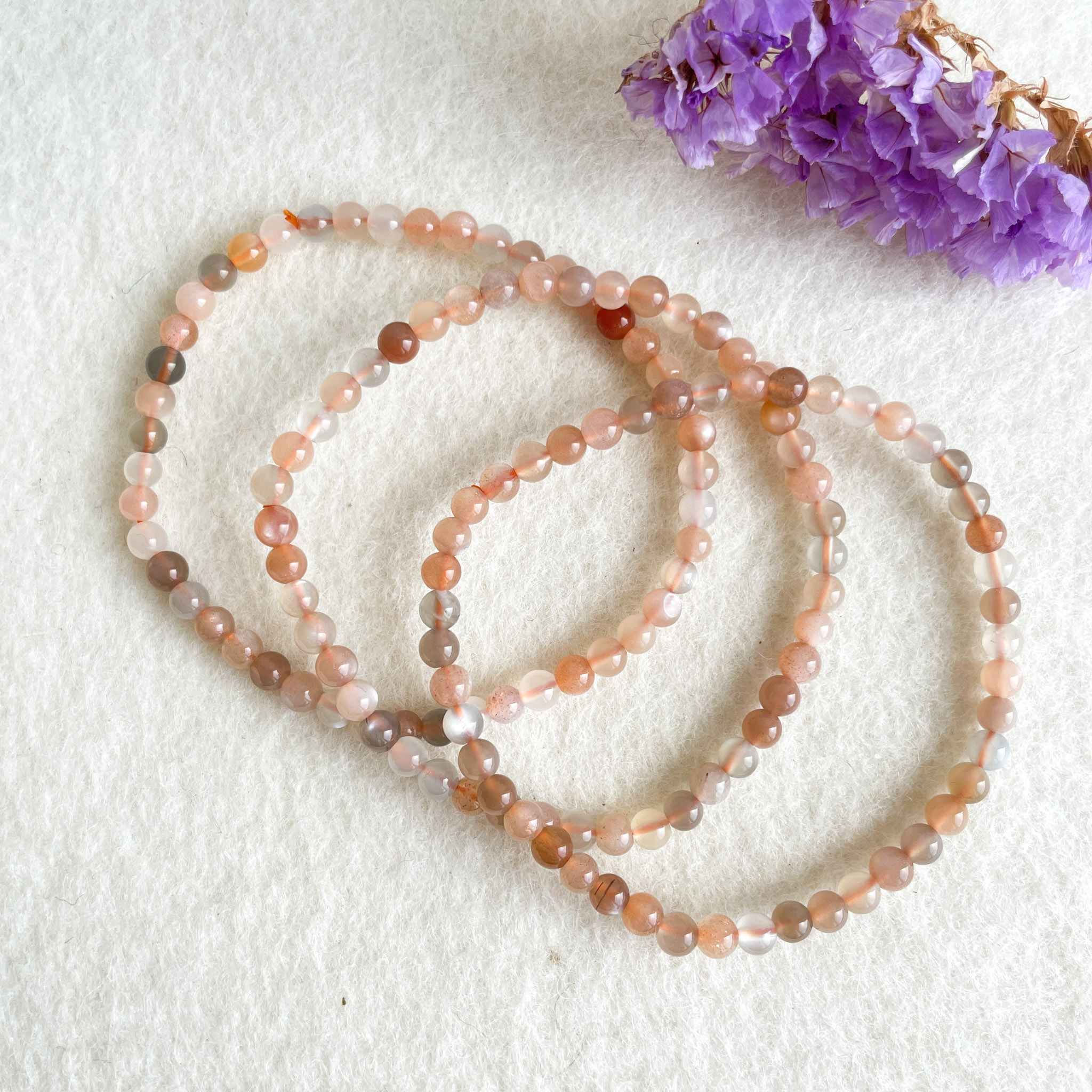Three strands of multicolored bead bracelets on a white textured background with a cluster of purple flowers partially visible in the corner.