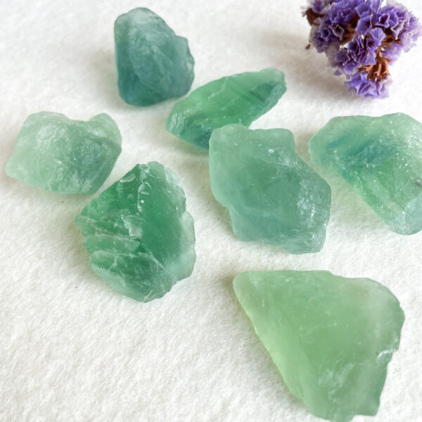 Several rough green gemstone crystals scattered on a white surface with a small purple dried flower in the top right corner.