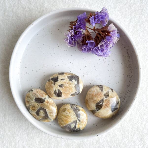 A white speckled ceramic bowl on a textured surface contains dried purple flowers and five rounded stones with natural beige and grey markings.