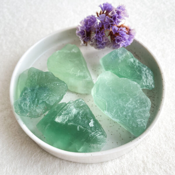 A ceramic plate containing rough green translucent gemstone chunks and decorated with a small bunch of purple flowers on a textured white surface.