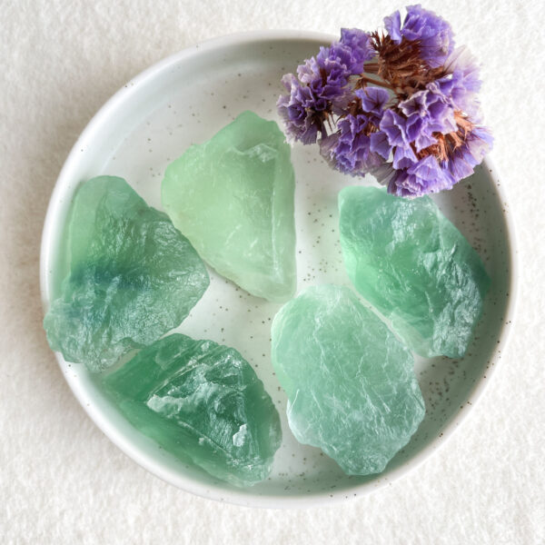 A white plate with speckles containing several translucent green gemstone pieces and a small bunch of dried purple flowers.