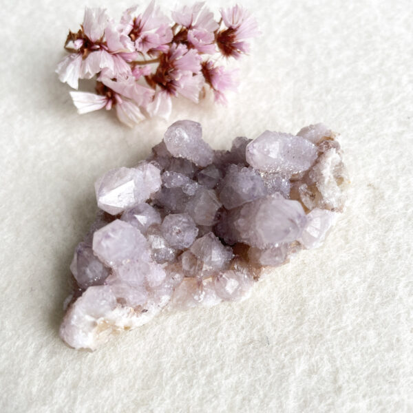 Alt text: A cluster of light purple amethyst crystals accompanied by a small bunch of pink flowers on a white textured background.