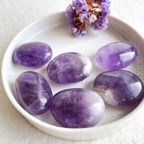 Several polished amethyst stones in a ceramic bowl with a dried purple flower on a textured white surface.