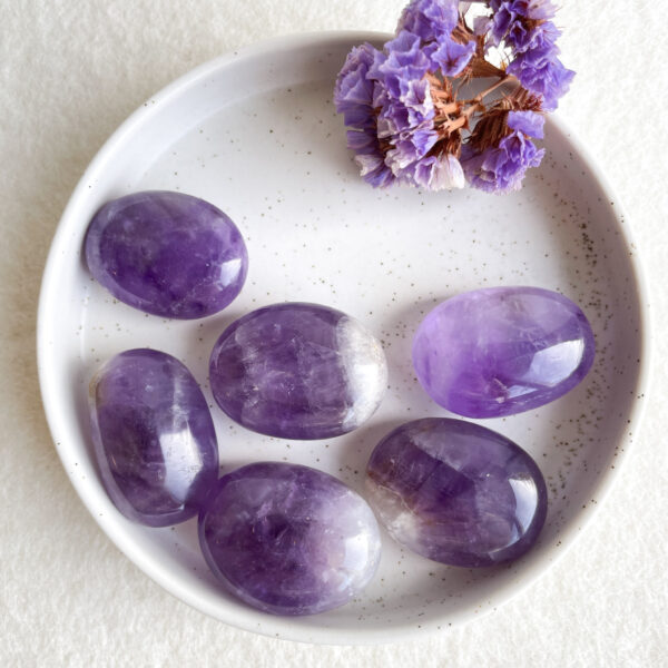 A white ceramic dish containing several polished amethyst stones and a small bunch of dried purple flowers on a textured white surface.