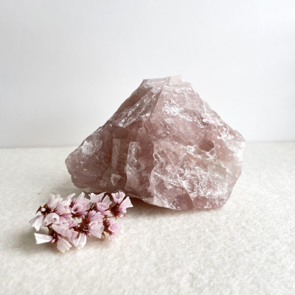 A large pink quartz crystal next to a small cluster of pink flowers on a white surface with a pale background.
