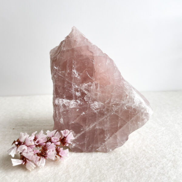 A large translucent pink crystal with rough edges sits on a white surface, accompanied by a small cluster of delicate pink flowers to its lower left. The background is plain and white, emphasizing the crystal's color and form.