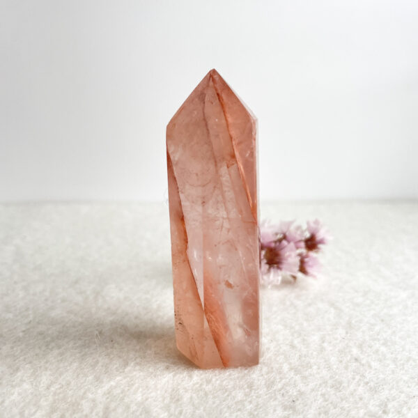 A translucent pink crystal tower with a faceted structure, displayed upright on a white surface with soft texture, with small pink flowers blurred in the background.