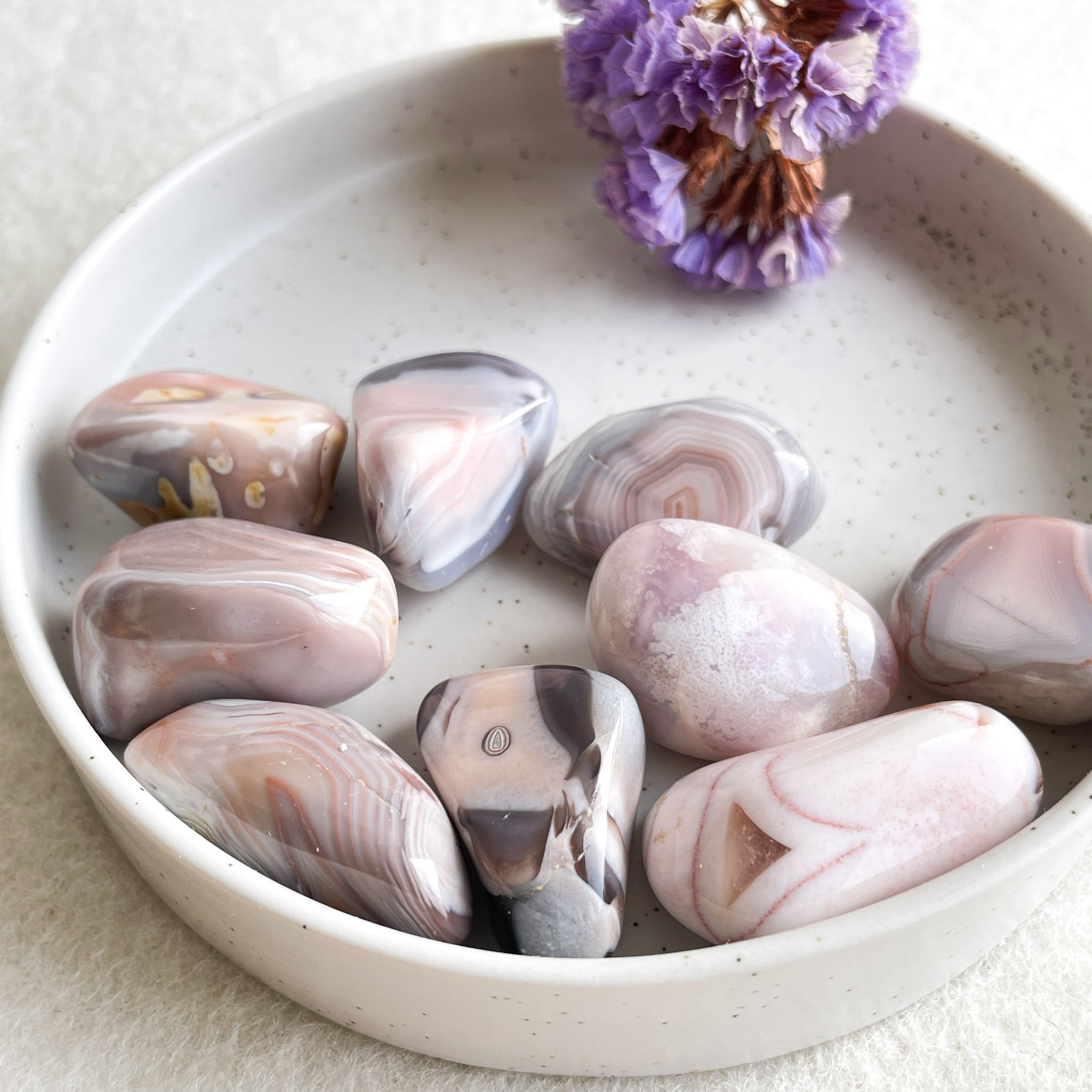 Polished agate stones in varying shades of pink and gray arranged in a white bowl beside a dried purple flower.