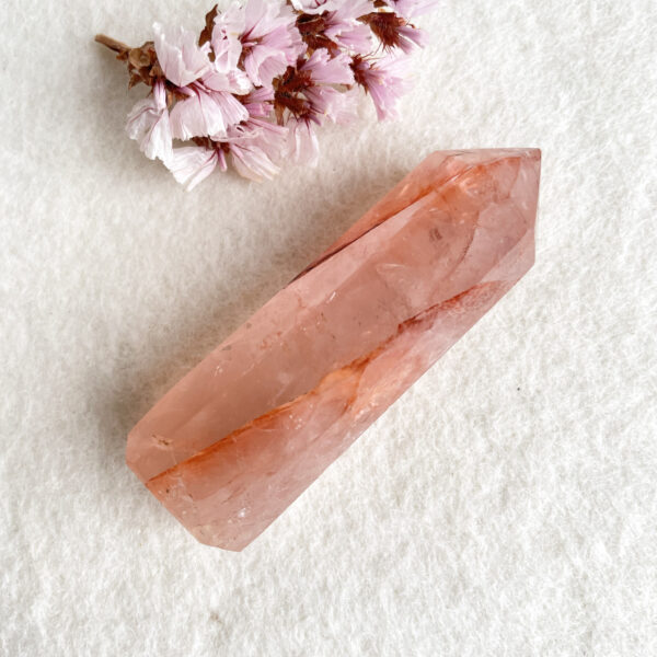 Alt text: A pink quartz crystal lays on a white textured background, accompanied by some wilted cherry blossoms to its left side.
