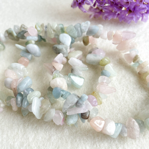 A close-up photo of pastel-colored gemstone chip bracelets on a soft, white background with a blurred purple flower in the distance.