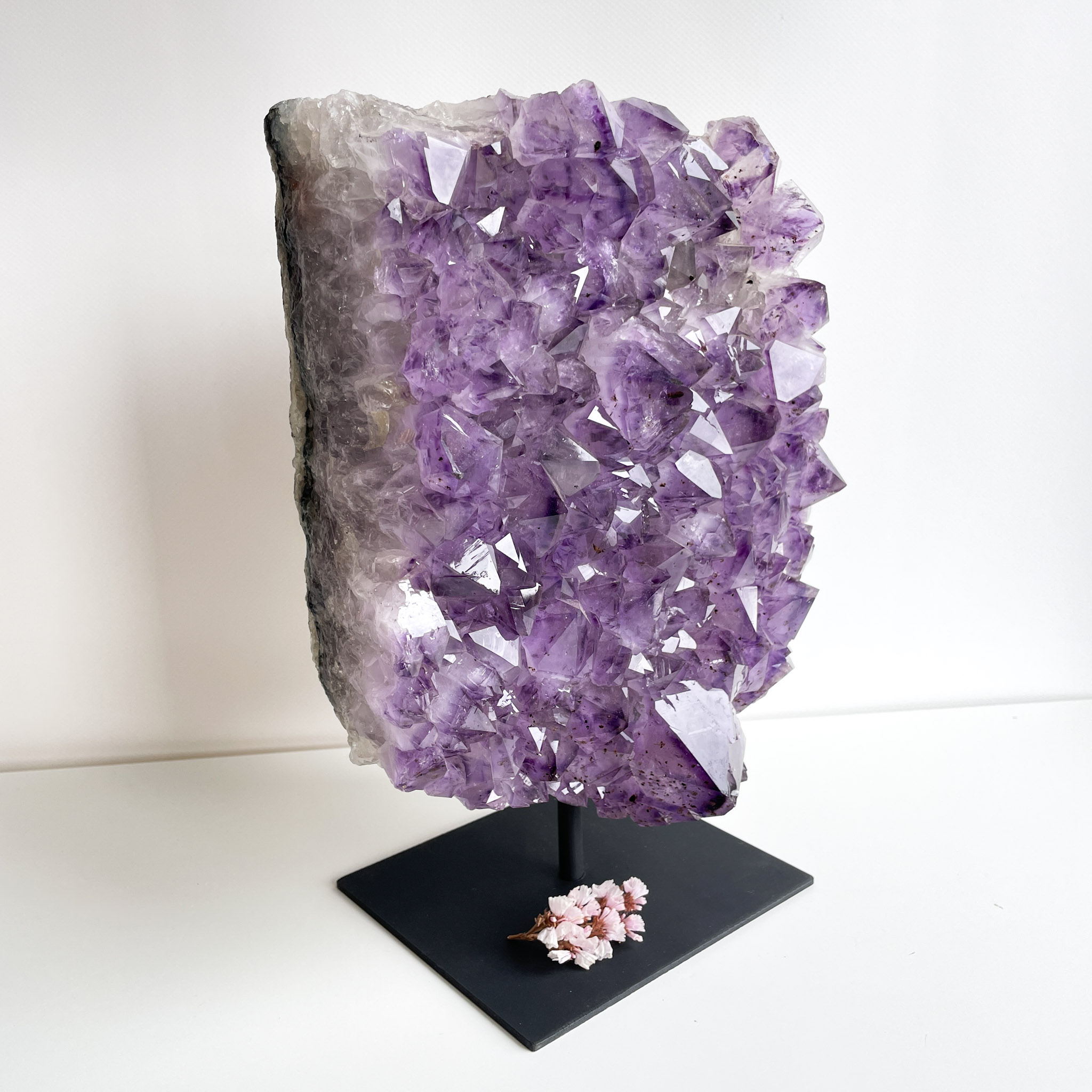 A large amethyst geode on a black stand with a cluster of pink flowers at its base, displayed against a white background.