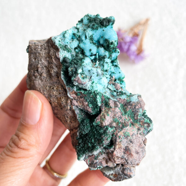 A hand holding a rough mineral specimen with vibrant green crystal formations on a rocky matrix.