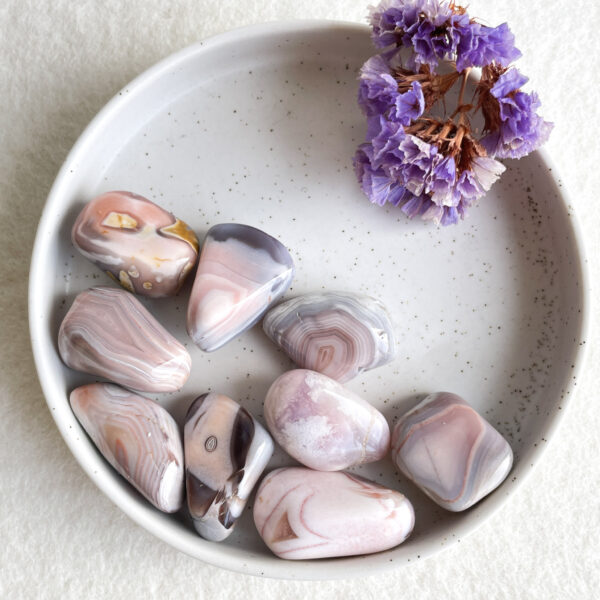 A collection of polished agate stones in shades of pink, white, and grey, displayed in a speckled ceramic bowl alongside a cluster of dried purple flowers.