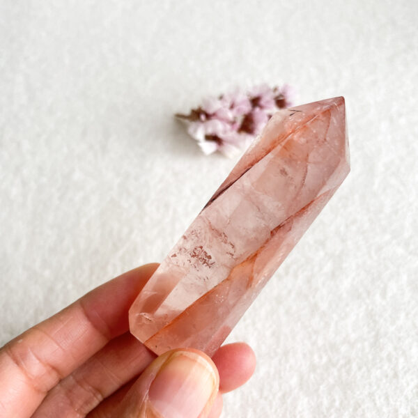 A person holds a translucent pink crystal wand with natural inclusions, with a blurred floral object in the background on a textured white surface.