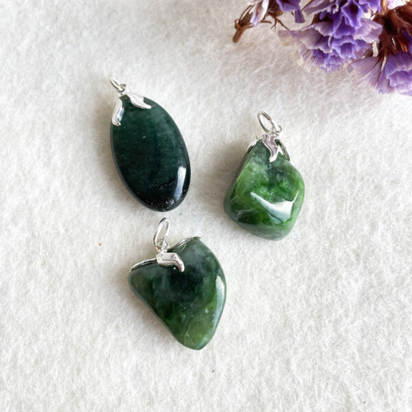 Three polished green stone pendants with silver bails on a white background, with the right side partially decorated with purple dried flowers.