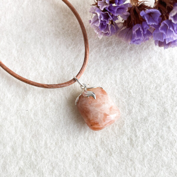 A leather necklace with a polished pink stone pendant on a white surface, partially surrounded by purple flowers.
