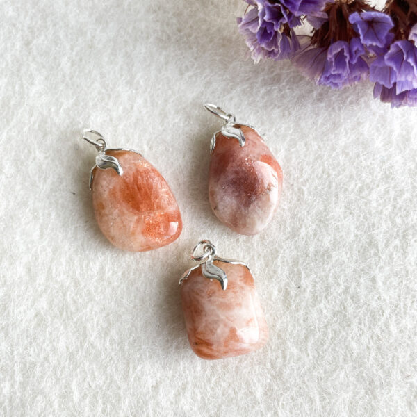 Three sunstone pendants with silver clasps on a white background, with purple flowers partially visible in the upper right corner.