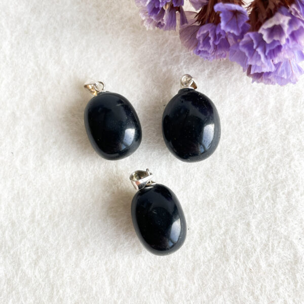 Three polished black stone pendants with silver mounting loops on a white textured surface, accompanied by purple flowers in the background.