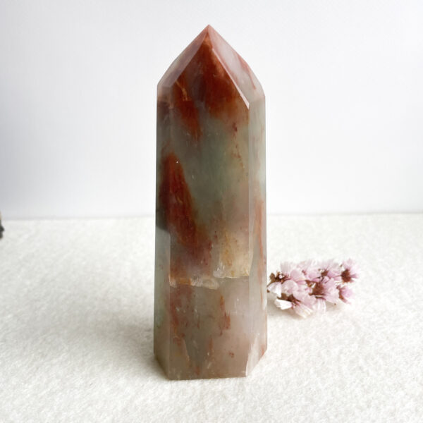 A translucent polished crystal tower with hues of red and orange on a textured white background, accompanied by a small cluster of pink flowers to the right.
