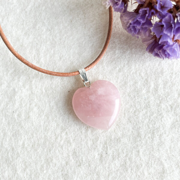 A pink heart-shaped pendant on a brown leather cord with a silver clasp lies on a white textured surface, with purple flowers partially visible in the corner.