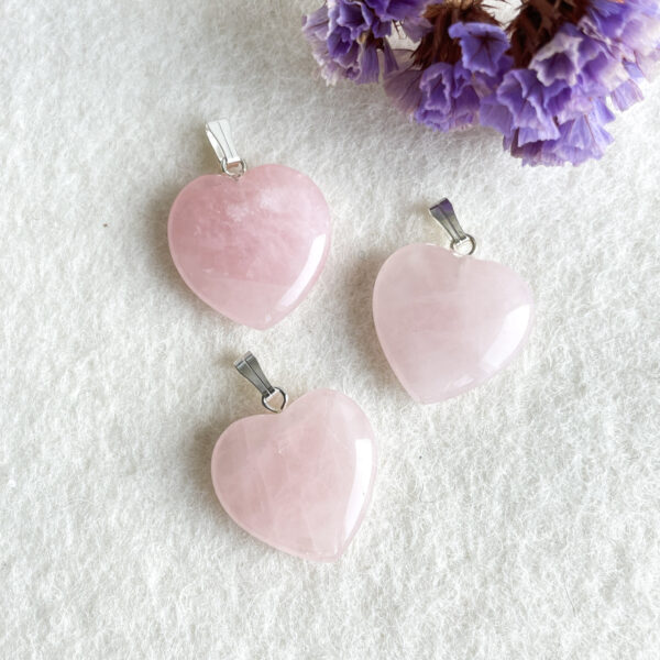 Three pink heart-shaped pendants with metallic loops for chains are laid out on a white fuzzy surface, with purple flowers in the top right corner.