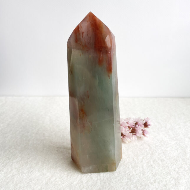 A polished multicolored crystal tower standing on a white surface with small pink flowers blurred in the background.